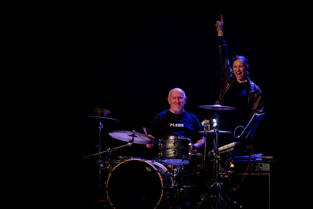 une comédienne derrière une personne jouant de la batterie
